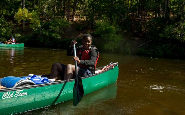 A person wearing a life jacket paddles a canoe on calm water near a tree-lined shore.